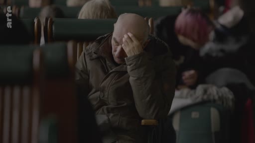 Un homme chauve, visiblement fatigué ou préoccupé, est assis dans un wagon de train, la main sur le front. Il est entouré d'autres passagers dans une ambiance sombre et calme.