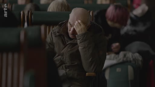 Un homme âgé, chauve, est assis dans un wagon de train, la main sur le front, semblant fatigué ou préoccupé. Il est entouré d'autres passagers dans un environnement de transport public.