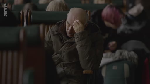 Un homme âgé au crâne dégarni, portant une veste sombre, est assis dans un wagon de train et semble préoccupé ou fatigué, la main sur le front. Il est entouré d'autres passagers dans une ambiance feutrée et sombre.