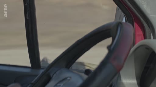 Vue subjective depuis l'intérieur d'un véhicule en mouvement, montrant le volant et le paysage extérieur dévasté, illustrant une situation de conflit ou de zone de guerre.