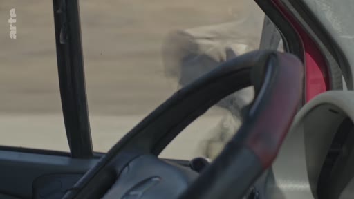 Vue subjective depuis le poste de conduite d'un véhicule traversant un paysage aride et poussiéreux, illustrant le déplacement dans une zone de conflit ou une région isolée.