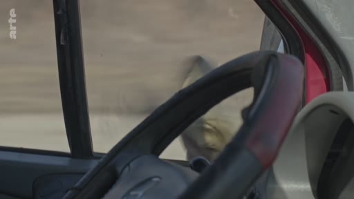 Vue subjective depuis le poste de conduite d'un véhicule circulant dans un paysage aride et désertique, évoquant une situation de reportage en zone de conflit ou de terrain difficile.