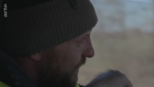 Un homme barbu portant un bonnet est filmé en gros plan alors qu'il conduit un véhicule, dans le cadre d'un reportage sur l'observation de zones dévastées.