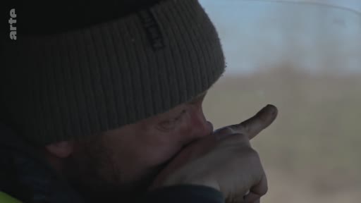 Un homme portant un bonnet sombre observe attentivement le paysage extérieur depuis l'intérieur d'un véhicule, illustrant une scène d'observation de la dévastation.
