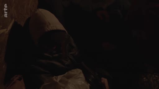 Un réfugié portant une capuche est assis dans l'obscurité à l'intérieur d'un véhicule, illustrant les conditions précaires du transport clandestin de migrants.