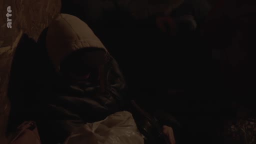 Une personne anonyme, le visage partiellement dissimulé par une capuche, est assise dans un environnement sombre, évoquant une situation de clandestinité ou de refuge.