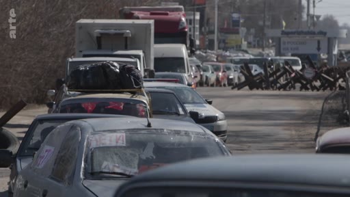 Une longue file de voitures chargées de bagages attend devant un barrage routier composé de hérissons tchèques, illustrant l'évacuation de civils dans une zone de conflit en Ukraine.