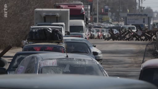 Une file de véhicules civils chargés de bagages attend à un point d'évacuation en Ukraine, bloquée par des herses métalliques anti-char sur la route. La scène illustre les difficultés rencontrées par les populations civiles lors des tentatives de fuite des zones de conflit.