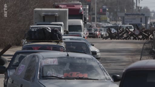 Une file de véhicules civils chargés de bagages attend à un point de contrôle avec des herses antichars à Zaporijia, en Ukraine, lors d'une opération d'évacuation. Le panneau indique 'Zaporizhzhia.city'.