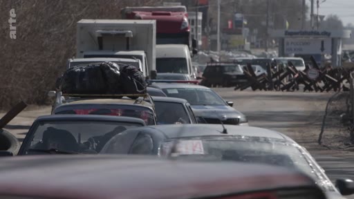 Une file de voitures chargées de bagages attend devant un barrage routier composé de hérissons tchèques, illustrant une situation d'évacuation de civils dans un contexte de conflit armé.
