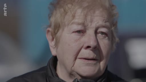 Une femme âgée s'exprime lors d'une interview, racontant son expérience personnelle liée à un conflit. Elle est filmée en gros plan, le visage marqué par l'émotion.