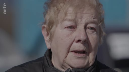 Une femme âgée s'exprime lors d'une interview, racontant son expérience de fuite face à un conflit. Elle porte une veste sombre et s'exprime avec émotion.