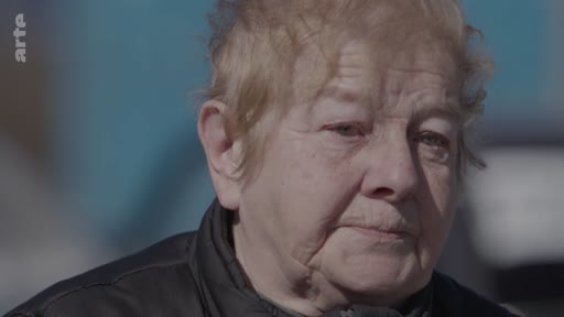 Une femme âgée s'exprime lors d'une interview, racontant son expérience personnelle liée à un conflit. Le plan serré met en avant son visage marqué par l'émotion.