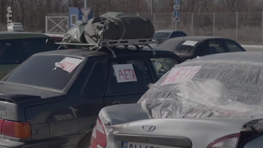 Des véhicules civils chargés de bagages et portant des panneaux « DÉTI » (enfants) en cyrillique sur leurs vitres, illustrant une situation d'évacuation de civils dans un contexte de conflit.