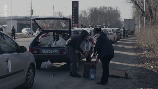 Des civils organisent l'évacuation de leurs biens en chargeant des sacs et des provisions dans le coffre d'une voiture, stationnée sur le bas-côté d'une route près d'une station-service, dans un contexte de crise humanitaire.