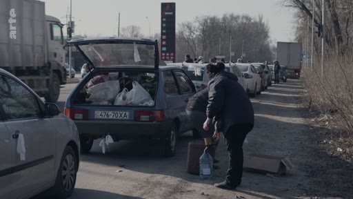 Des civils ukrainiens chargent des sacs et des effets personnels dans le coffre d'une voiture sur le bord d'une route, dans un contexte d'évacuation. On aperçoit une file de véhicules et une station-service en arrière-plan.