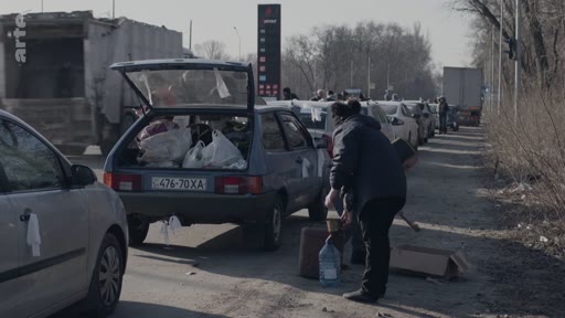 Des civils chargent des effets personnels dans leurs voitures sur le bord d'une route, illustrant une situation d'évacuation d'urgence dans un contexte de conflit. On aperçoit une file de véhicules et des personnes s'organisant pour quitter la zone.