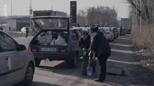Des civils ukrainiens chargent des bagages et des bidons d'eau dans le coffre d'une voiture sur une route encombrée, illustrant l'exode des populations fuyant les zones de conflit.