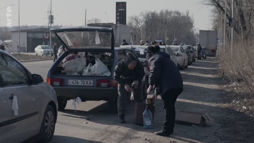 Des civils ukrainiens chargent des sacs et des provisions dans le coffre d'une voiture sur le bord d'une route lors d'une évacuation. La scène montre des personnes en train de préparer leur véhicule pour fuir une zone de conflit, avec d'autres voitures stationnées en file indienne à l'arrière-plan.