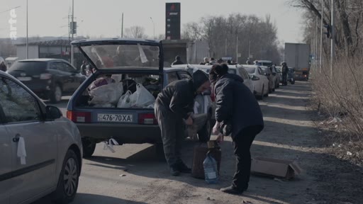 Des civils organisent le chargement de leurs effets personnels et de provisions dans le coffre d'une voiture sur le bord d'une route, dans un contexte d'évacuation de population. La scène montre des personnes en plein préparatifs de départ, avec des véhicules chargés en arrière-plan.
