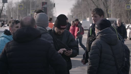 Un groupe d'hommes, dont certains portent des bonnets, se rassemble et discute au bord d'une route urbaine. L'un des hommes consulte son téléphone portable au milieu du groupe.