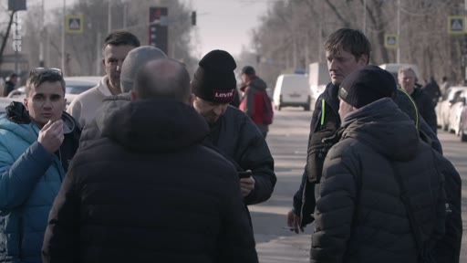 Un groupe d'hommes, dont certains portent des bonnets, est rassemblé au bord d'une route urbaine pour discuter. La scène se déroule en extérieur, dans un contexte de rassemblement informel.