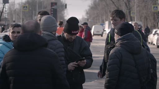Un groupe d'hommes, dont certains portent des bonnets, se rassemble et discute au bord d'une route urbaine. L'un des hommes consulte son téléphone portable pendant que les autres semblent échanger des informations dans un contexte de manifestation ou de rassemblement public.