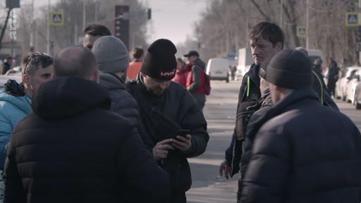 Un groupe d'hommes, dont certains portent des bonnets, se rassemble et discute au bord d'une route urbaine. L'un des hommes consulte son téléphone portable au milieu du groupe.