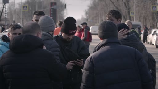 Un groupe de personnes, dont certaines portent des bonnets, se rassemble au bord d'une route en zone urbaine. Un homme au centre consulte son téléphone portable.