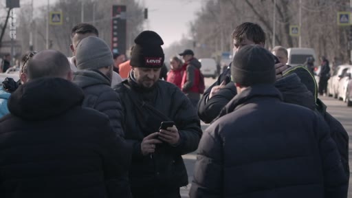 Un groupe d'hommes, dont certains portent des bonnets et des vestes d'hiver, se rassemble sur le bord d'une route en zone urbaine. L'un des hommes consulte son téléphone portable au milieu du groupe.