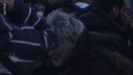Un homme âgé aux cheveux gris et à la barbe courte est filmé en gros plan au milieu d'une foule, dans une ambiance sombre et urbaine.