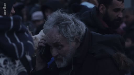 Un homme âgé avec une barbe grise téléphone dans une foule, probablement lors d'un déplacement de réfugiés ou d'une situation de crise humanitaire.
