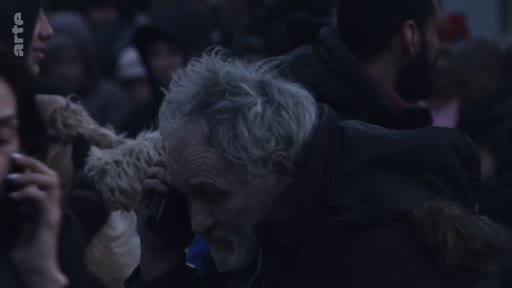 Un homme âgé aux cheveux gris téléphone dans une foule dense, probablement lors d'un déplacement de réfugiés ou d'une situation de crise humanitaire. L'ambiance est sombre et tendue, avec plusieurs personnes autour utilisant également leur téléphone.