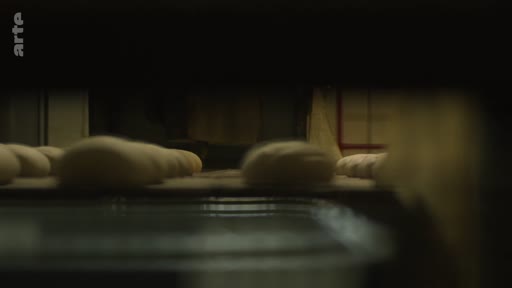 Des pâtons de pain crus sont transportés sur un tapis roulant dans une boulangerie industrielle située près de la ligne de front. Cette image illustre la production alimentaire en zone de conflit.