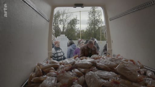 Des habitants font la queue pour recevoir des sacs de pain distribués depuis l'arrière d'un camion de livraison, illustrant une situation d'aide humanitaire ou de distribution alimentaire.