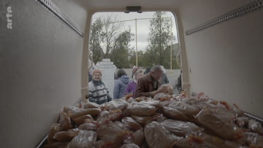Des habitants font la queue pour recevoir des sacs de nourriture distribués depuis l'arrière d'un camion dans un contexte d'aide humanitaire ou alimentaire. La scène montre une distribution de vivres à une foule de personnes dans un espace extérieur.