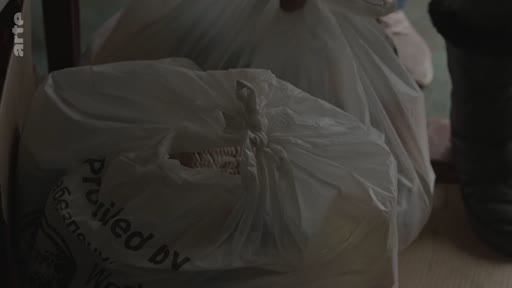 Des sacs en plastique blanc contenant de l'aide humanitaire sont empilés lors d'une opération de distribution. Le texte 'Provided by' est visible sur l'un des sacs.
