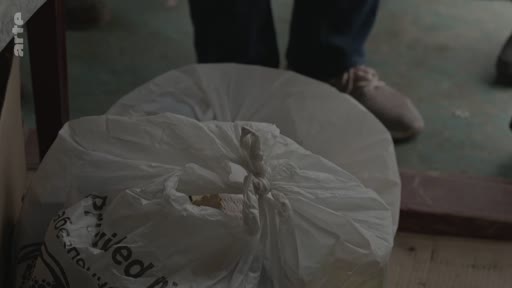 Un sac en plastique blanc contenant de l'aide humanitaire est posé au sol lors d'une opération de distribution. On aperçoit les pieds d'une personne à proximité.