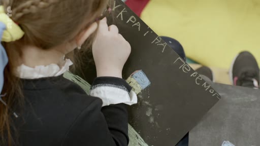 Une jeune enfant dessine sur une feuille de papier noir portant l'inscription manuscrite en ukrainien « УКРАЇНА ПЕРЕМОГ » (Ukraine victoire), illustrant le soutien des enfants ukrainiens à leur pays.
