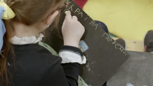 Une jeune fille écrit à la craie sur une ardoise noire le message « УКРАЇНА ПЕРЕМОЖЕ » (L'Ukraine gagnera) lors d'une activité de dessin. Cette scène illustre le soutien des enfants ukrainiens à leur pays dans le contexte du conflit actuel.