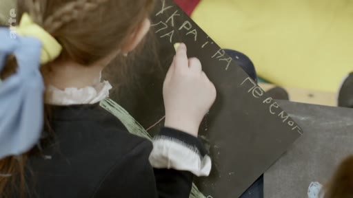 Une enfant écrit en ukrainien « УКРАЇНА ПЕРЕМОГ » (Ukraine victoire) à la craie sur un tableau noir, illustrant le soutien des jeunes générations ukrainiennes pendant le conflit.