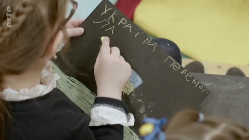 Une jeune enfant écrit à la craie « УКРАЇНА ПЕРЕМОГЛА » (L'Ukraine a gagné) sur une feuille de papier noir, illustrant le soutien des enfants ukrainiens face à la guerre.