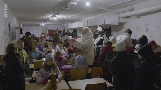 Des élèves et des enseignants se trouvent dans un abri anti-aérien souterrain aménagé au sein d'une école, probablement en Ukraine, en raison du conflit en cours. La pièce est bondée, avec des enfants assis à des pupitres scolaires, illustrant les conditions de vie précaires des civils pendant la guerre.