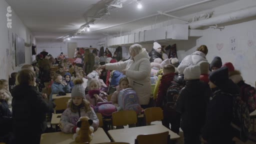 Des enfants et des adultes sont rassemblés dans un abri anti-aérien souterrain, aménagé dans une salle de classe, pour se protéger lors d'un conflit. La scène montre des élèves assis à leurs pupitres dans un environnement confiné, illustrant les conditions de vie des civils en temps de guerre.