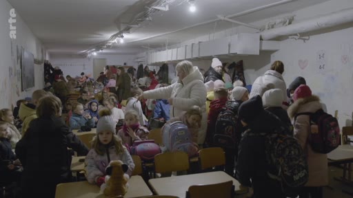Des élèves et des enseignants se trouvent dans un abri anti-aérien souterrain, aménagé dans les sous-sols d'une école, pour se protéger des bombardements. La scène montre une atmosphère de tension et de résilience alors que les enfants attendent dans une salle de classe improvisée.