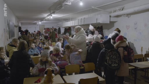 Des élèves et des enseignants se trouvent dans un abri anti-aérien souterrain aménagé au sein d'une école, probablement en Ukraine, pour se protéger des bombardements. La scène montre une foule d'enfants avec des sacs à dos dans un espace confiné et faiblement éclairé, illustrant les conditions de vie en temps de guerre.