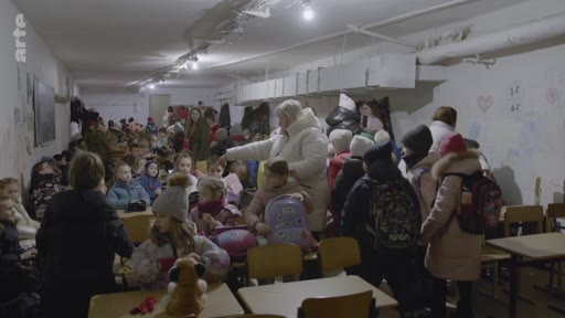 Des enfants et des enseignants se trouvent dans un abri anti-aérien souterrain aménagé dans une école, probablement en Ukraine, en raison du conflit en cours. La pièce est bondée d'élèves portant des sacs à dos, illustrant les conditions de scolarité en temps de guerre.