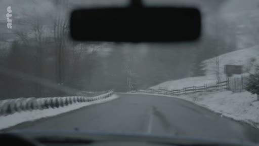Vue subjective depuis le poste de conduite d'un véhicule circulant sur une route de montagne couverte de neige, illustrant les conditions de circulation hivernales.