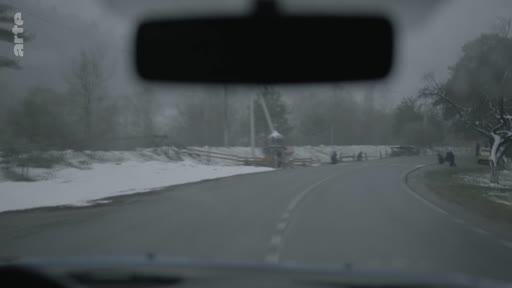 Vue subjective depuis le pare-brise d'un véhicule circulant sur une route de campagne bordée de neige, illustrant un trajet hivernal dans un village.
