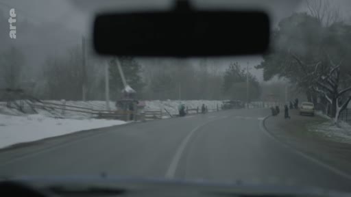 Vue depuis l'intérieur d'une voiture circulant sur une route bordée de neige dans un village rural, sous un ciel gris et hivernal.